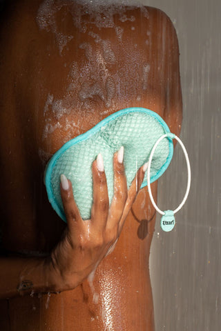 A close-up of a person using a turquoise Uzari exfoliating sponge on their wet skin while showering, with water streaming down.