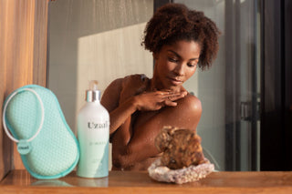 A woman enjoying a shower, gently touching her shoulder while water runs down. In the foreground, a Uzari U-Wash body cleanser and a turquoise exfoliating sponge are placed on a wooden surface beside a decorative stone.
