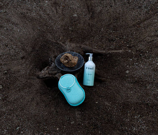 : A bottle of Uzari U-Wash body cleanser and a turquoise exfoliating sponge placed on dark sand next to a decorative stone on driftwood.