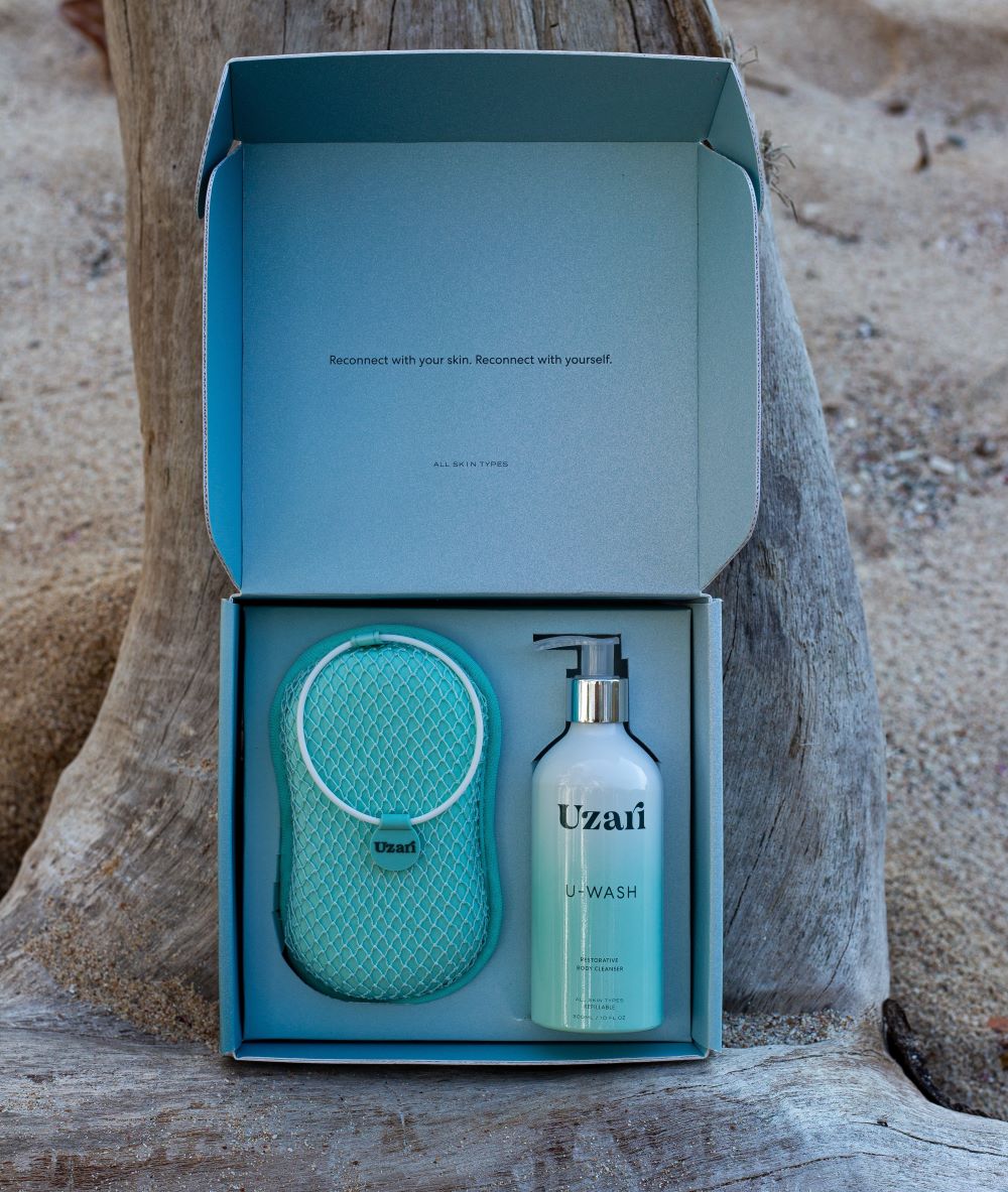 An open turquoise gift box displayed outdoors on a tree trunk, containing a Uzari U-Wash body cleanser bottle and a turquoise exfoliating sponge. 