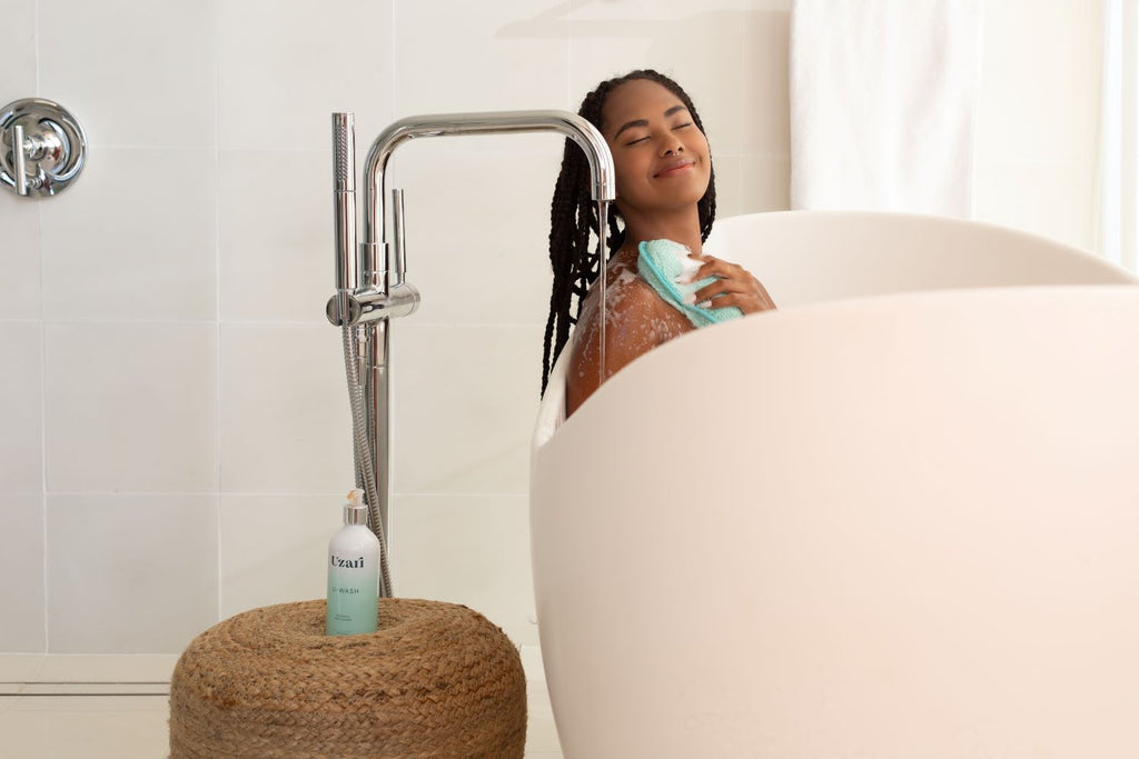 A woman relaxing in a white bathtub, smiling while using a turquoise exfoliating sponge with Uzari U-Wash body cleanser. The bottle of cleanser is placed on a woven stool next to the tub in a modern bathroom setting.