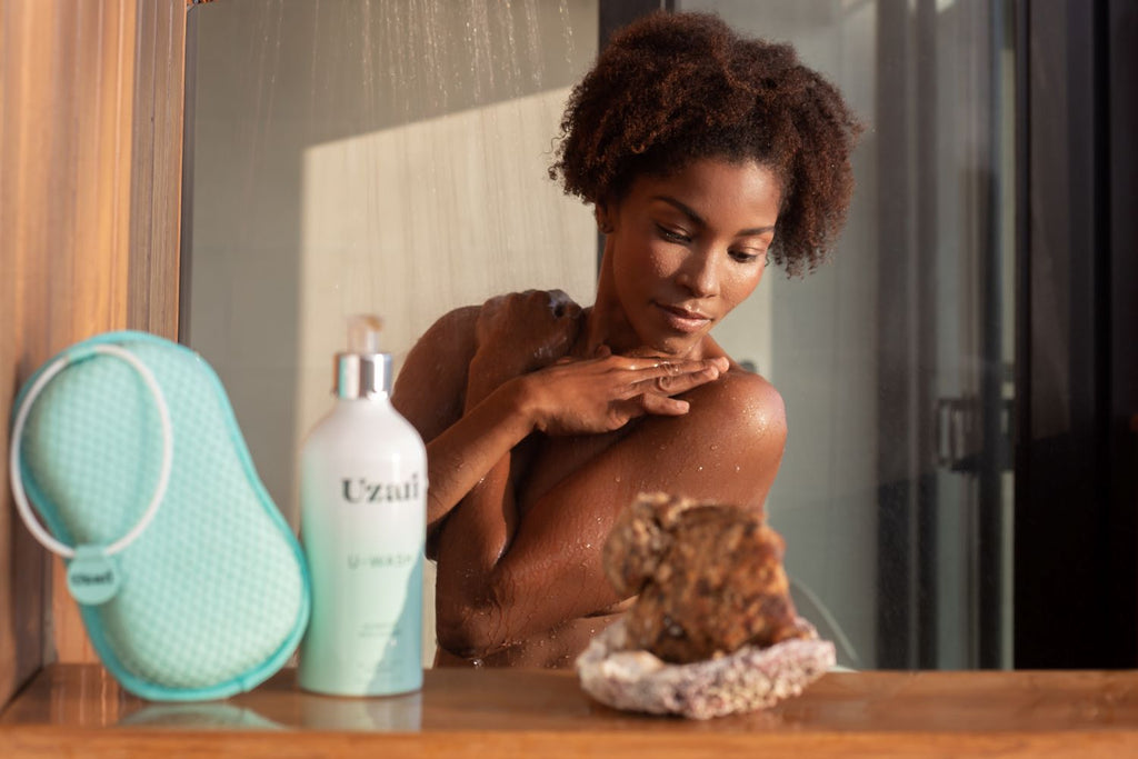 A woman enjoying a shower, gently touching her shoulder while water runs down. In the foreground, a Uzari U-Wash body cleanser and a turquoise exfoliating sponge are placed on a wooden surface beside a decorative stone.