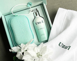 Uzai skincare product set with bottle, pouch, and towel on a gray surface with white flowers.