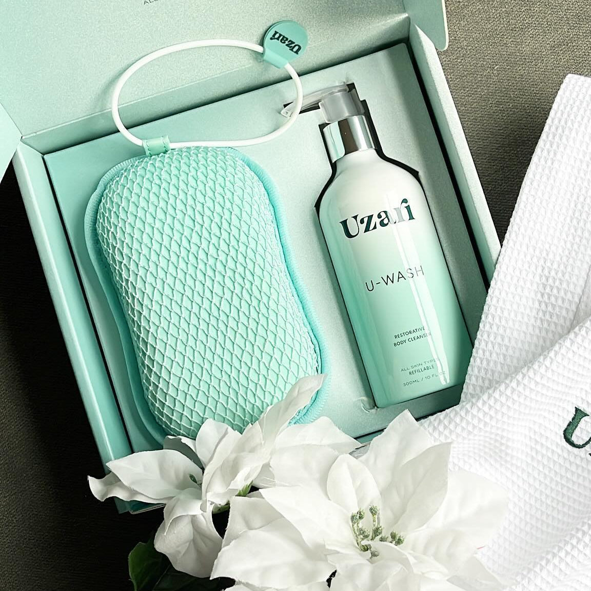 Uzai skincare product set with bottle, pouch, and towel on a gray surface with white flowers.