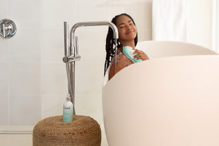 A woman relaxing in a white bathtub, smiling while using a turquoise exfoliating sponge with Uzari U-Wash body cleanser. The bottle of cleanser is placed on a woven stool next to the tub in a modern bathroom setting.